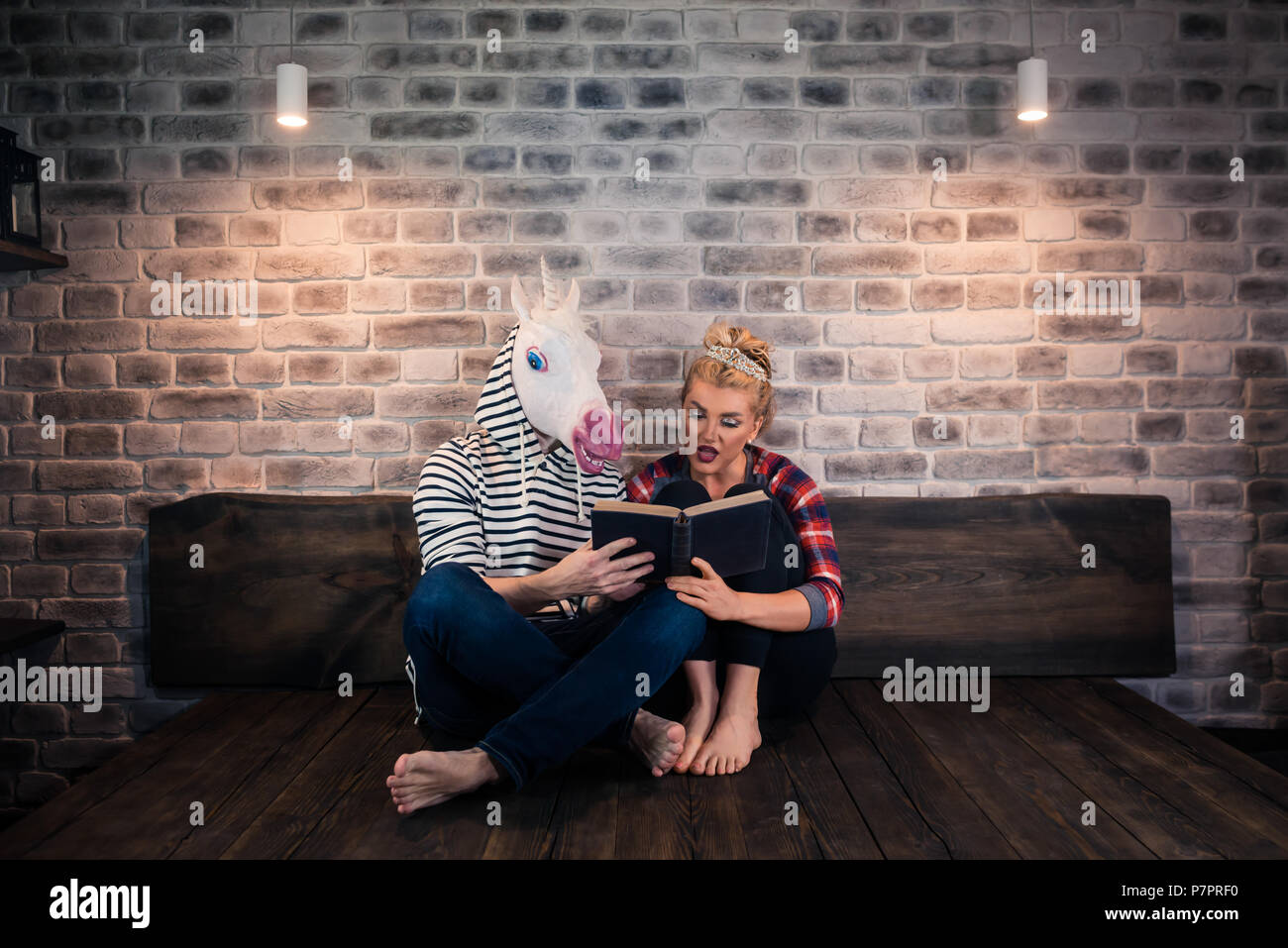 Ungewöhnliche Paar ein Buch lesen in stilvolle Apartment. Schöne Mädchen sitzt mit lustigen Mann in komischen Maske. Junge Frau mit freakigen Freund zu Hause Stockfoto