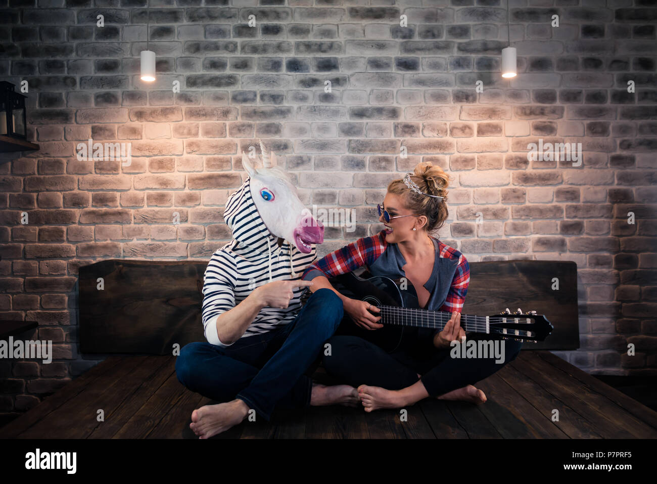 Blonde Mädchen spielen Musik auf Gitarre für freaky Mann in komischen Maske. Ungewöhnliche Paar verbringen viel Zeit gemeinsam in der stilvollen Apartments. Einhorn mit Freundin Stockfoto