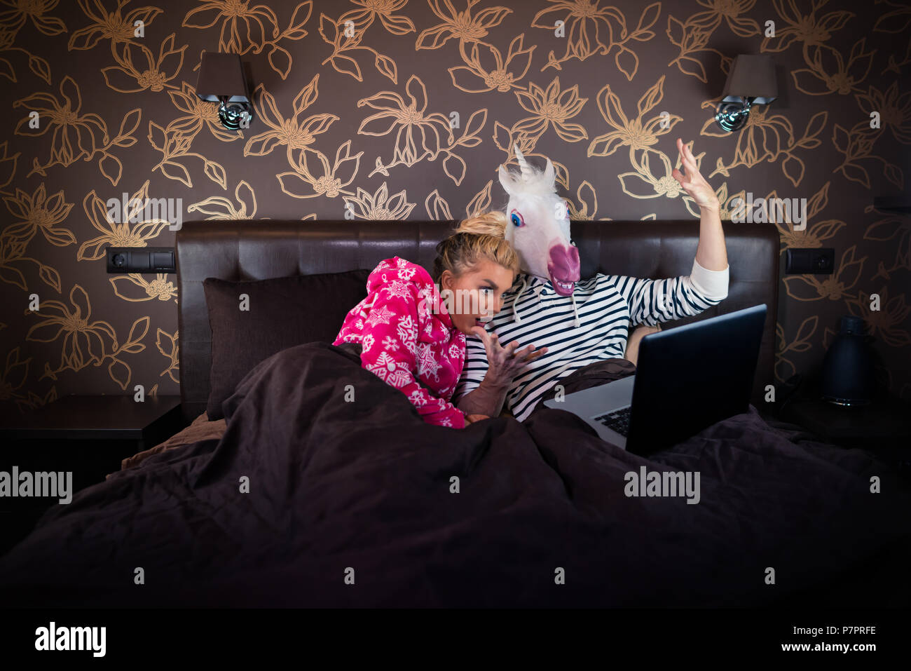 Überrascht Mädchen ist auf dem Bett liegend mit lustigen Freund in komischen Maske. Ungewöhnliche Paar schaut Film auf Laptop in der stilvollen Schlafzimmer. Stockfoto