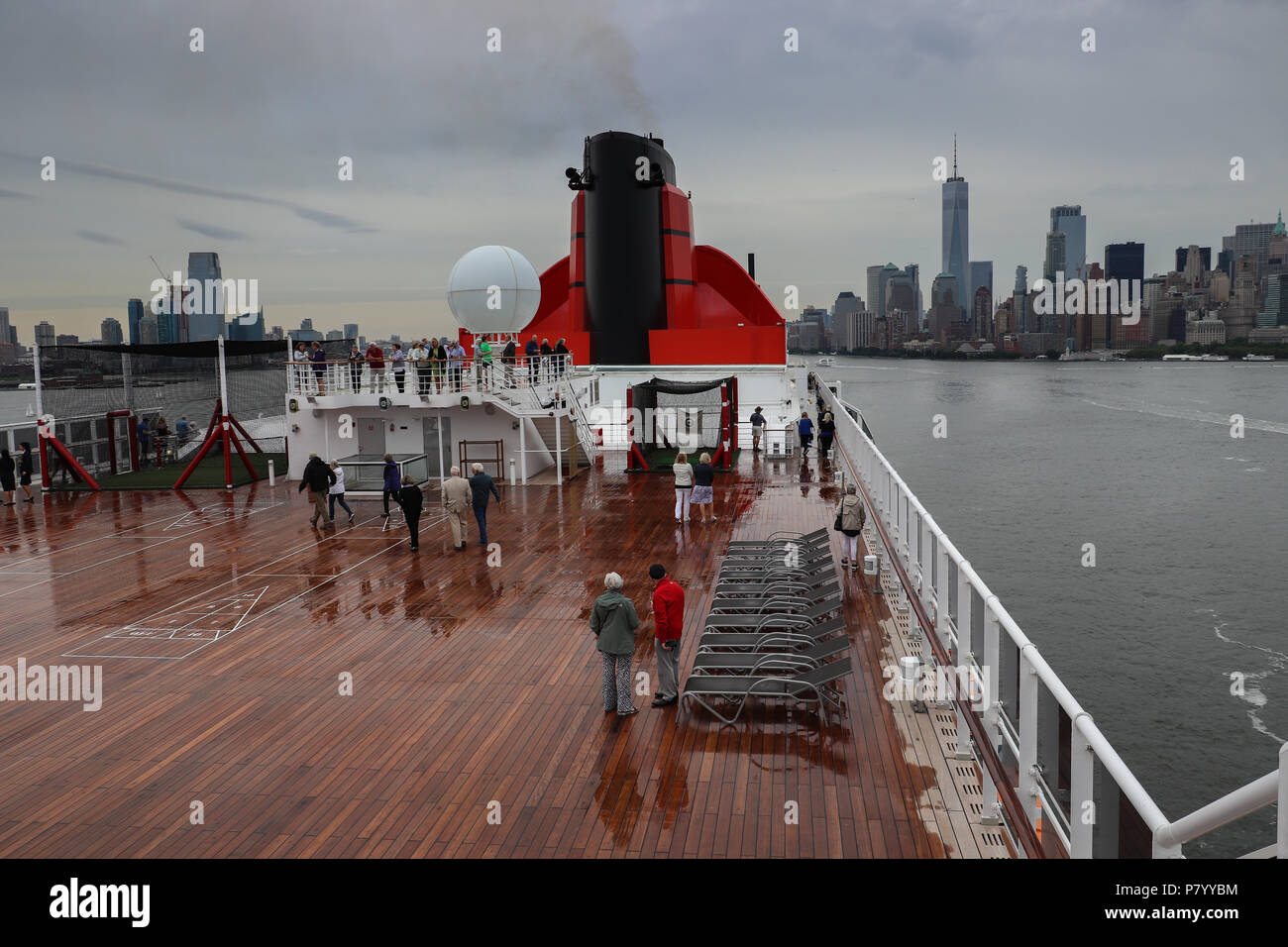 Die Passagiere sehen Sie sich die "Queen Mary 2" den Hafen von New York aus einem nassen Deck verlassen an einem regnerischen Abend, die Skyline von New York City in der Ferne sichtbar. Stockfoto