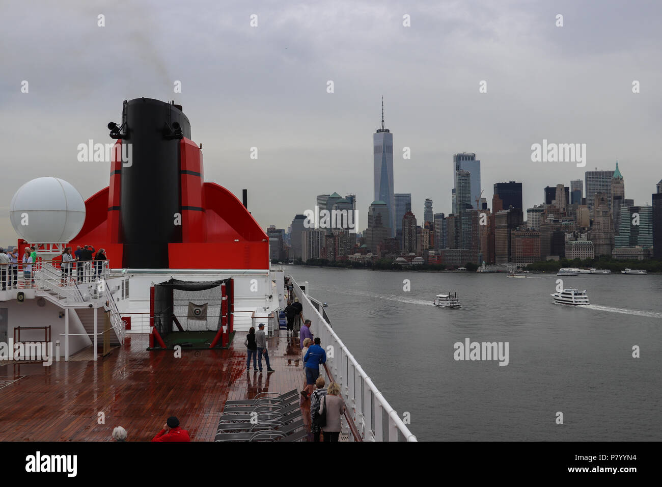 Die "Queen Mary 2" fährt von New York Hafen an einem regnerischen Tag, mit der Skyline der Stadt und das One World Trade Center im Hintergrund sichtbar; zwei Boote. Stockfoto