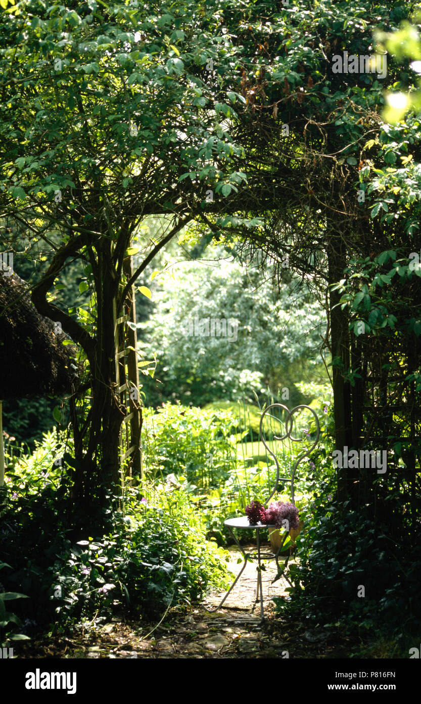 Reich verzierten schmiedeeisernen Stuhl gegen Gitter Bogen mit grünen Kletterpflanzen und Blick auf Country Garden Stockfoto