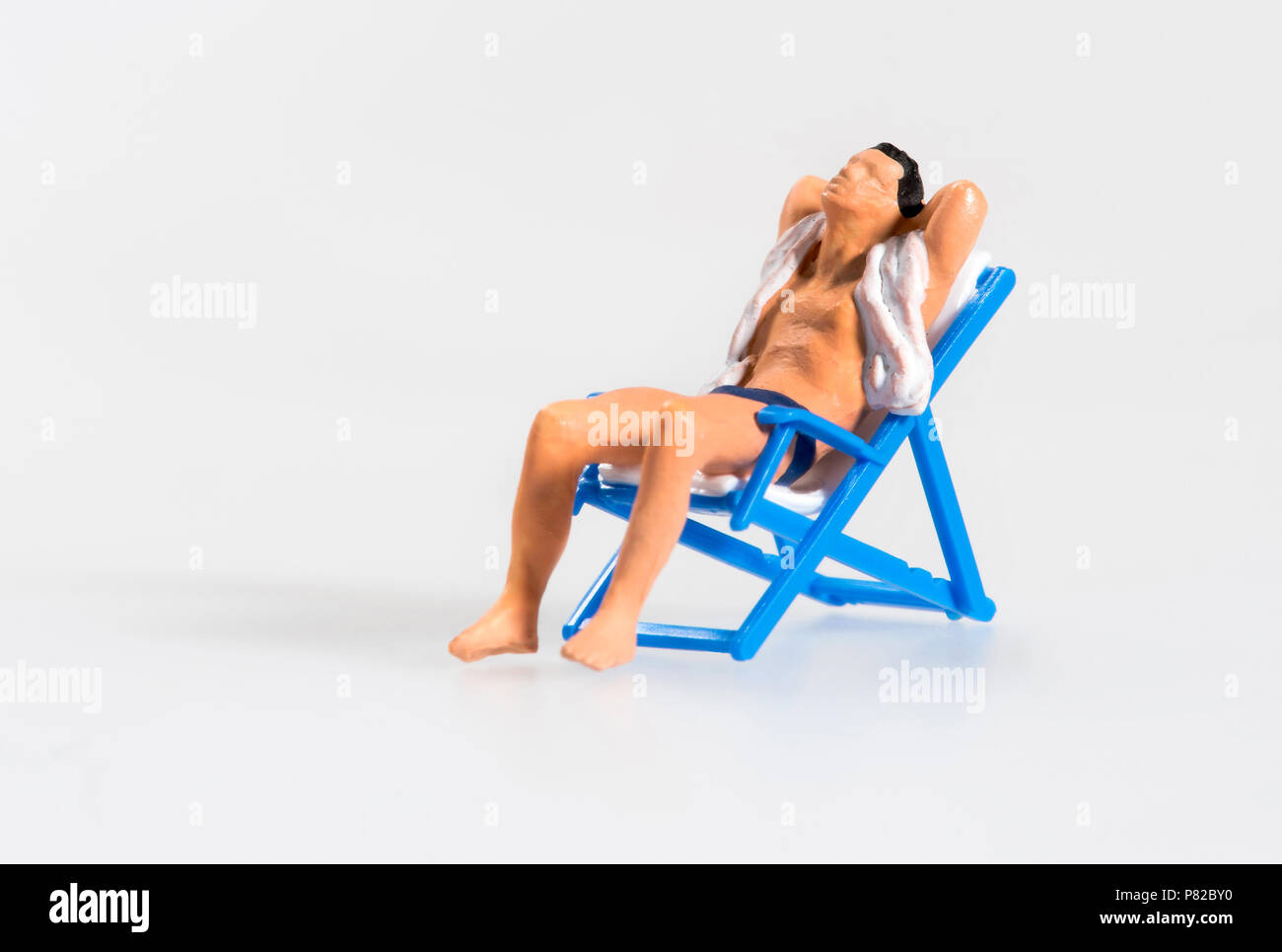 Miniatur Mann in T-Shirt und Badehose entspannenden Sonnenbad auf einer klappbaren blau Liegestuhl Sessel mit seiner Hand in den Nacken konzeptionelle eines Sommers Stockfoto