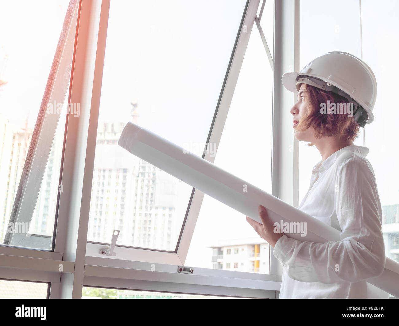 Schöne asiatische Frau Ingenieur curly kurze Haare tragen weiße Schutzhelm und weißes Hemd holding Blaupause in der Nähe der Fenster aus Glas mit Aufbau Stockfoto