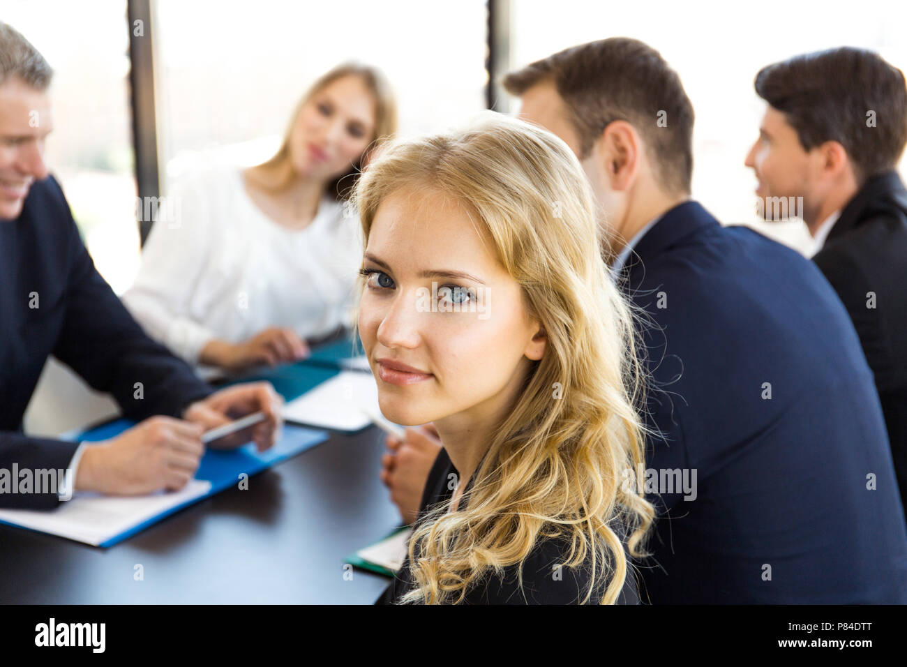 Die Leute sitzen rund um Tisch, Reden und Arbeiten mit Dokumenten, den Schwerpunkt auf die junge Frau an der Kamera auf der Suche Stockfoto