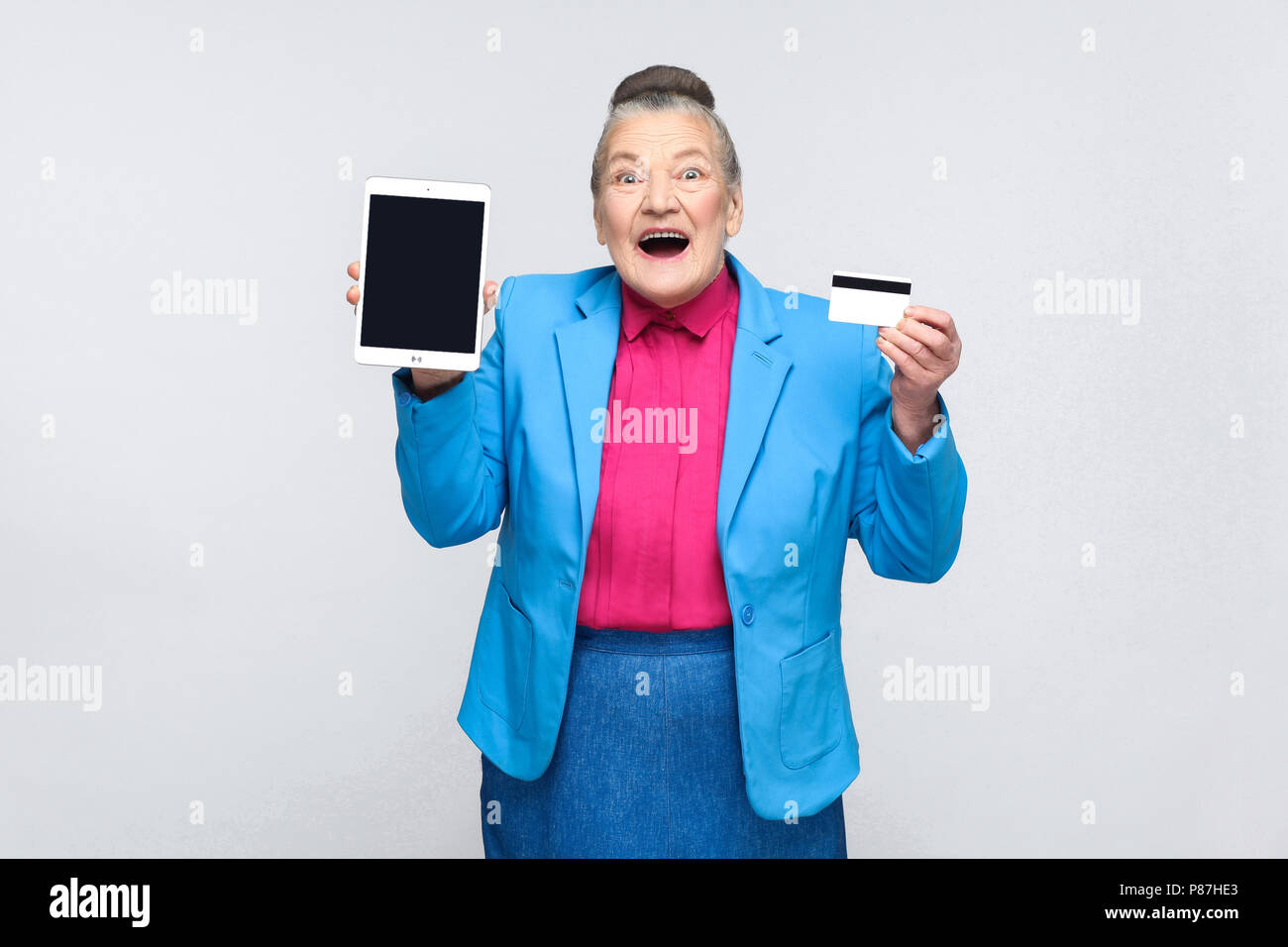 Gealterte Frau zeigen tablet Bildschirm und Kreditkarte und Suchen von der Kamera begeistert. Großmutter im blauen Anzug mit gesammelten graue Haare bun Frisur. Ich Stockfoto
