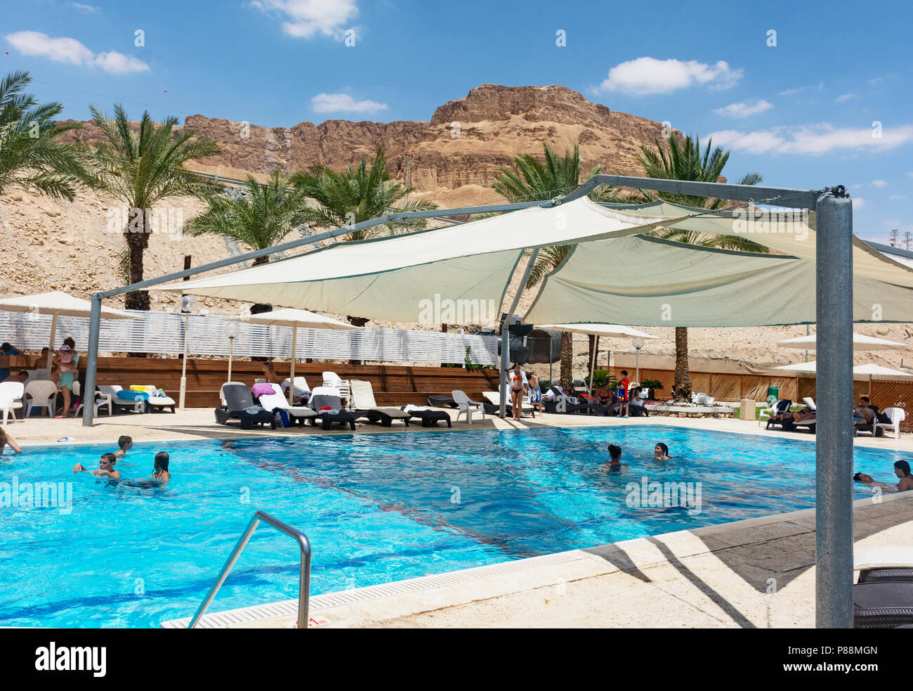 Touristen genießen einen teilweise schattigen Außenpool in einem Resorthotel in ein Bokek am Toten Meer mit Wüstenbergen und blauem Himmel Stockfoto