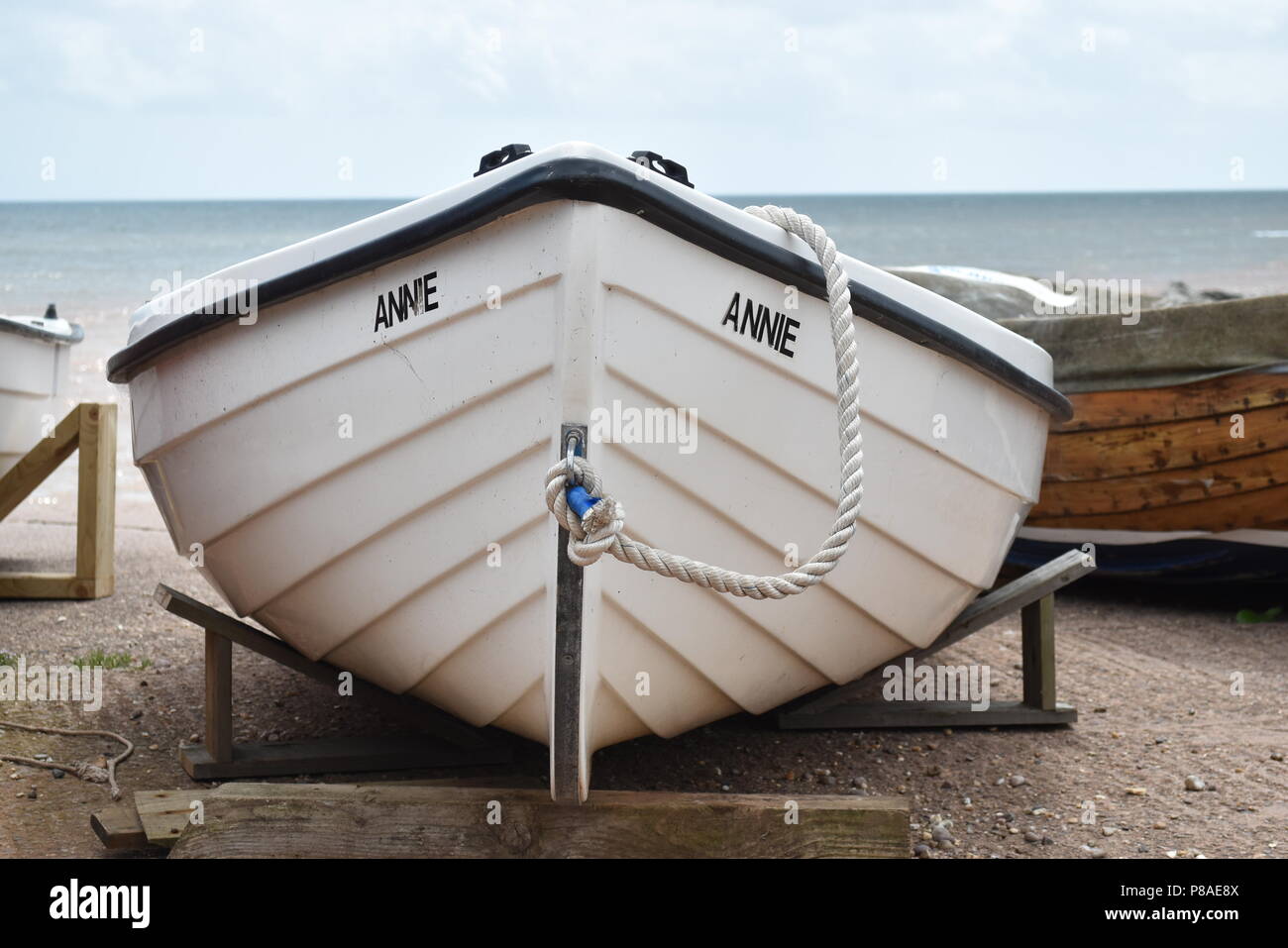 Der Bug eines kleinen weißen Boot namens Annie ruht auf unterstützt in der Nähe der Küste mit dem Meer im Hintergrund in Honiton, Devon, England Stockfoto