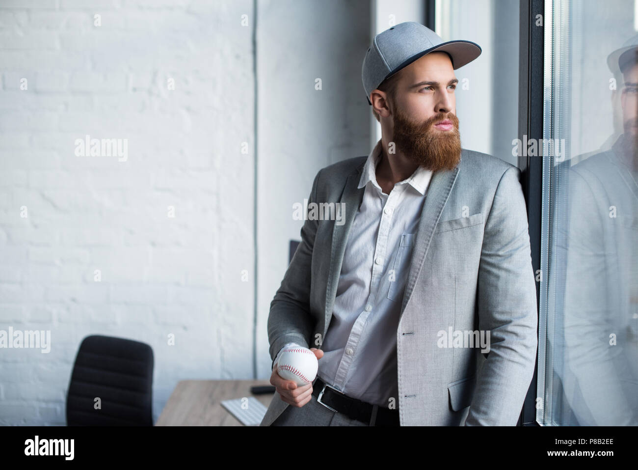 Bärtige Geschäftsmann im Baseball Kappe am Fenster schauen und Holding baseball ball Stockfoto