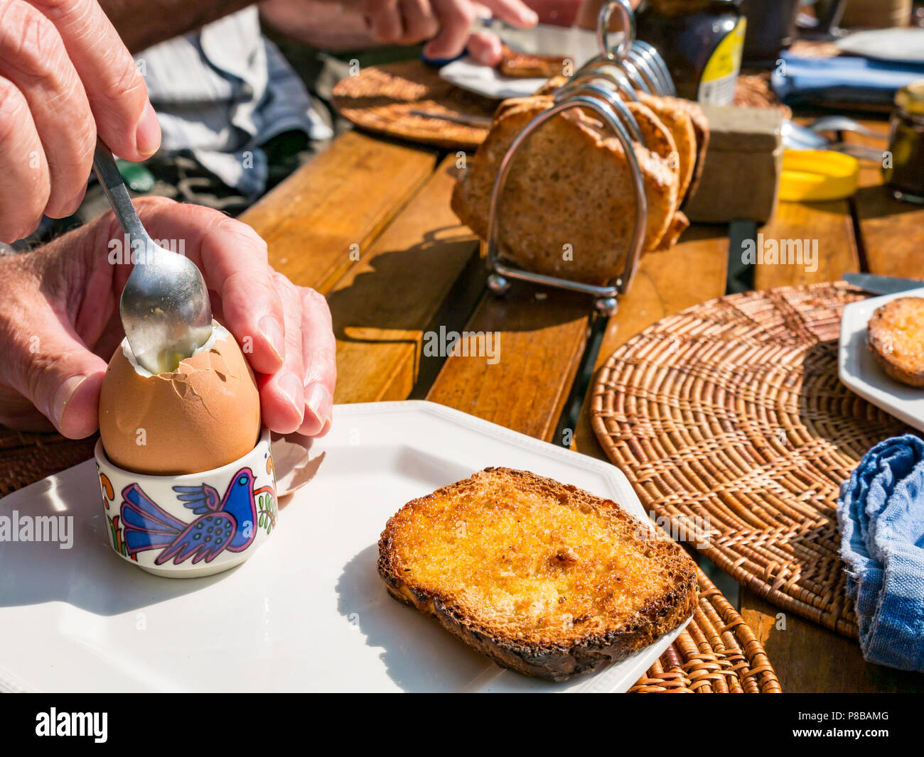 Mann, der gekochtes Ei in einer Eierschale mit Toast auf einem Holzgartentisch im Freien mit Toastablage bei Sonnenschein zum Frühstück isst Stockfoto