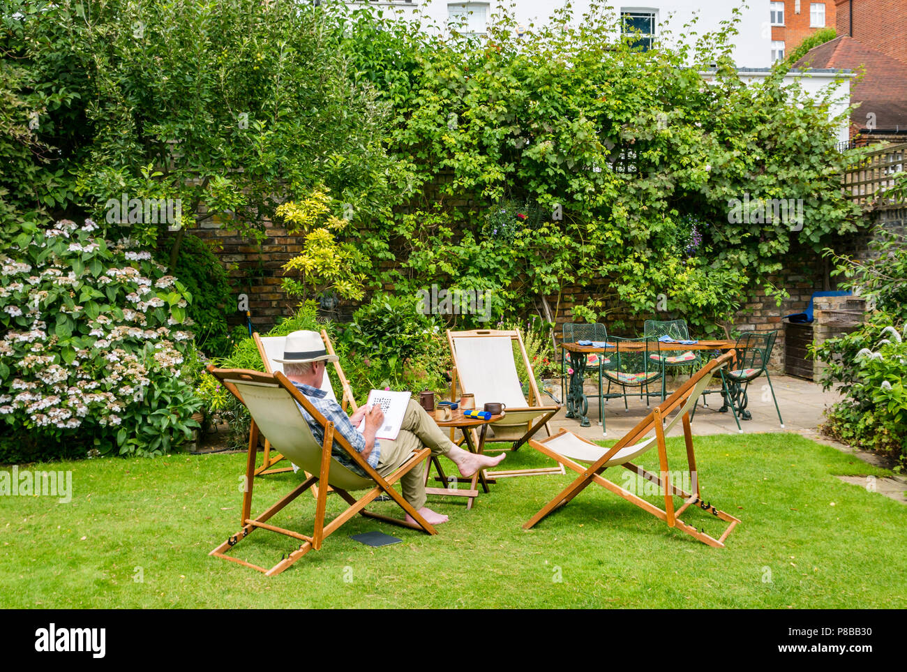 Ältere Mann, der Panama Hut Lösung Kreuzworträtsel im Liegestuhl sitzen im großen Garten in Sommerhitze, London, England, Großbritannien Stockfoto