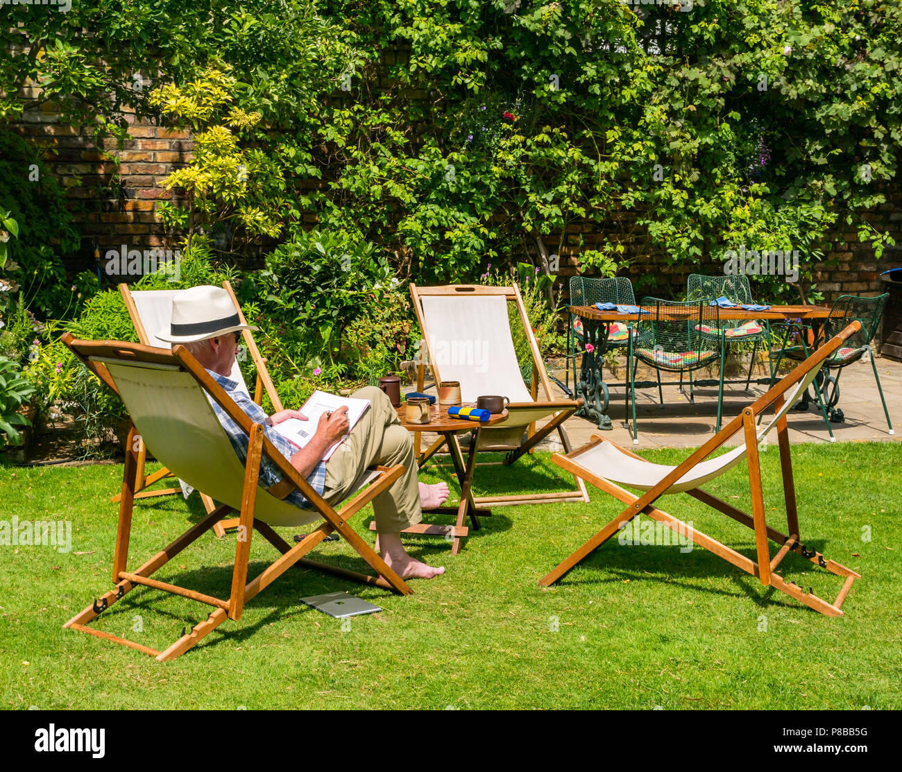 Ältere Mann, der Panama Hut Lösung Kreuzworträtsel im Liegestuhl sitzen im großen Garten in Sommerhitze, London, England, Großbritannien Stockfoto