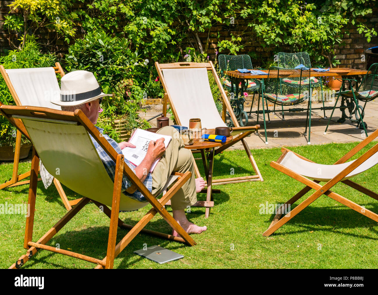 Ältere Mann, der Panama Hut Lösung Kreuzworträtsel im Liegestuhl sitzen im großen Garten in Sommerhitze, London, England, Großbritannien Stockfoto