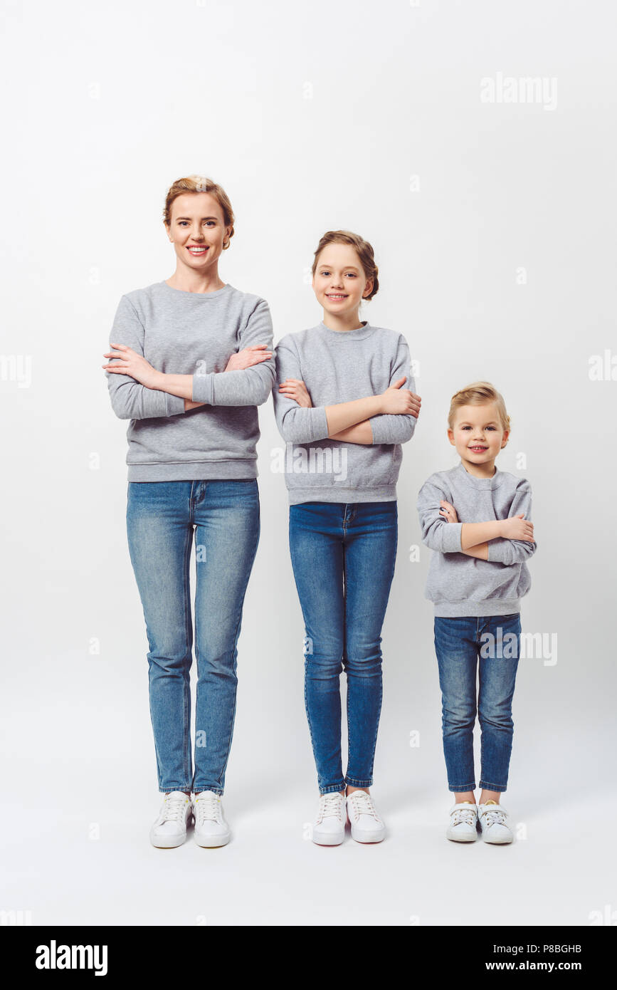 Lächelnde Mutter und Töchter der verschiedenen Generationen in ähnlicher Kleidung mit verschränkten Armen auf Grau isoliert Stockfoto