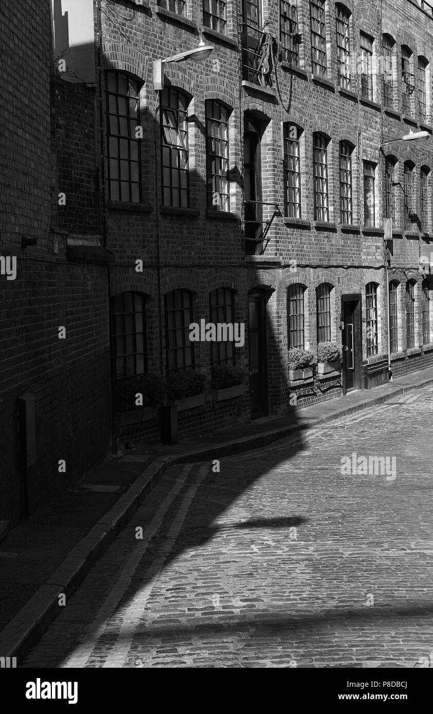 Schwarz-weiß-Bild der Mauer Lagerhallen und gepflasterte Strasse mit pflastersteine im East End von London. Schatten auf der Wand des Gebäudes und der Stockfoto