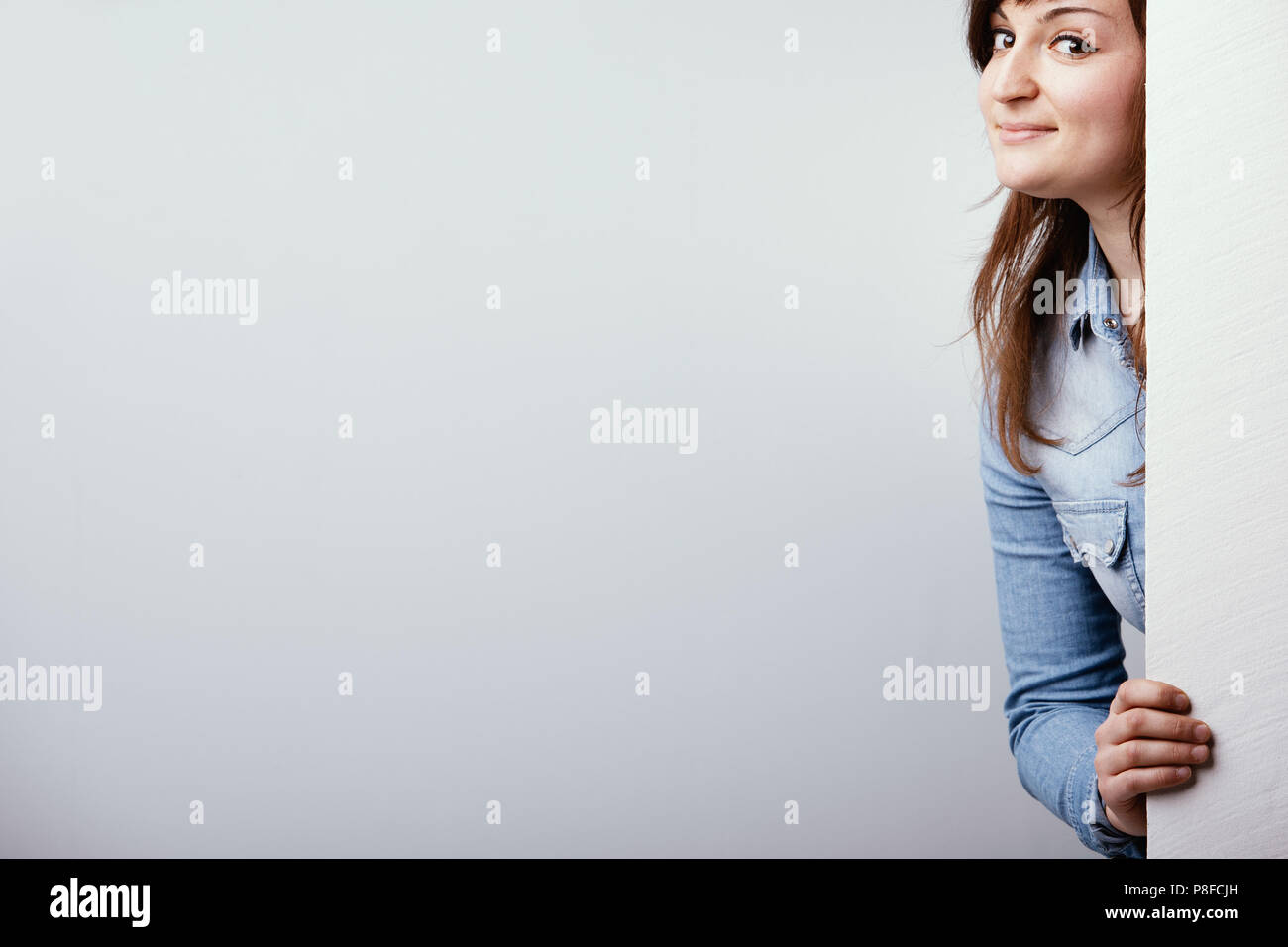 Freche junge Frau Peering um ein weißes Schild mit einem schelmischen Grinsen über einem weißen studio Hintergrund mit Kopie Raum Stockfoto