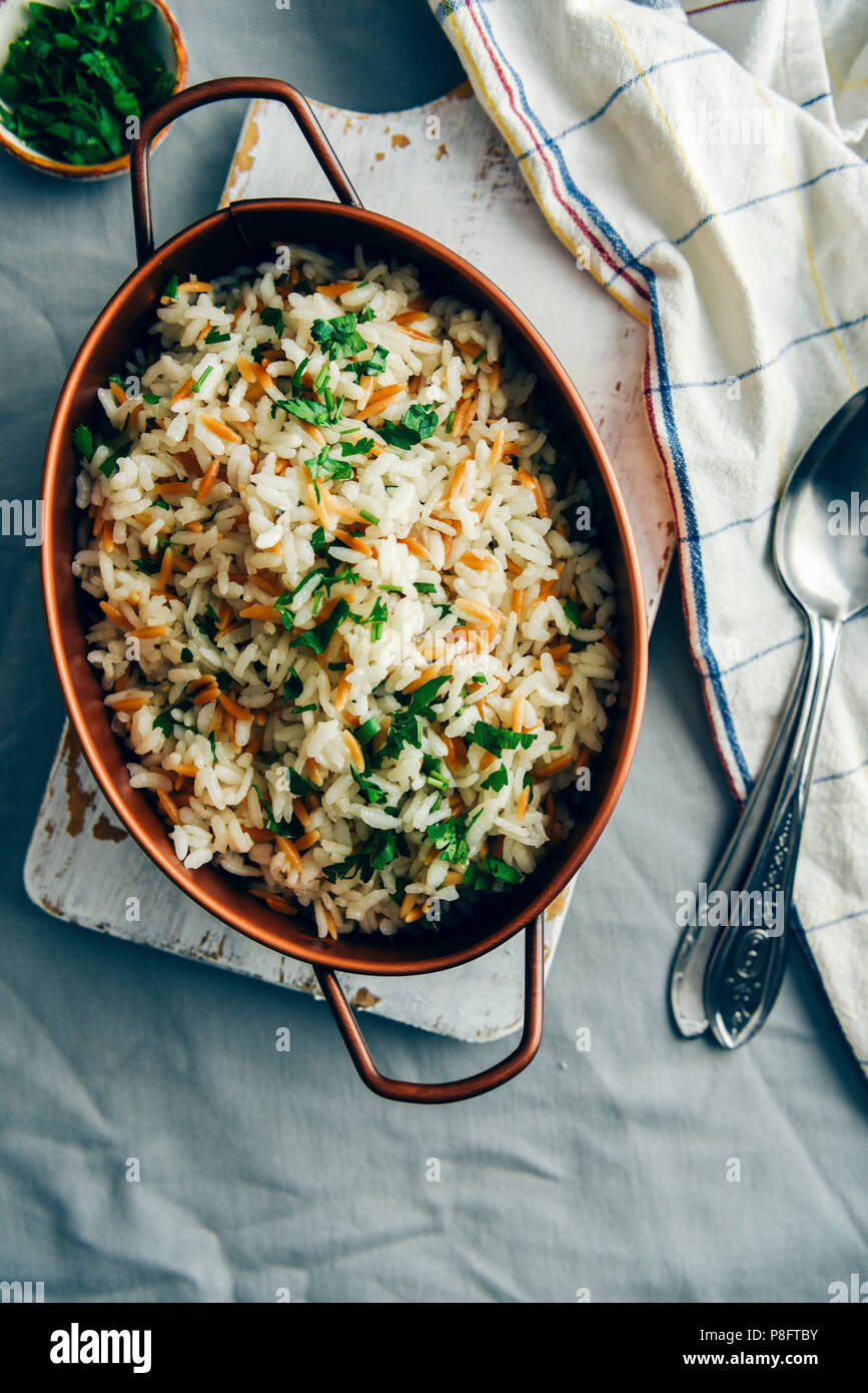 Gebratener Reis Pilav mit orzo in einem Kupfer serviert pan auf eine weiße Tafel. Löffel, weisse Serviette und eine kleine Schüssel mit gehackter Petersilie begleiten. Stockfoto