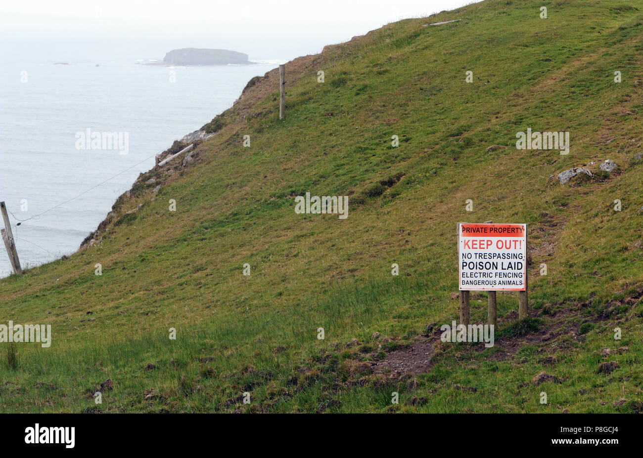 Ein hysterischer Schild mit der Aufschrift "PRIVATEIGENTUM", "KEEP OUT!", "KEINE ÜBERTRETUNG", "GIFT LAID", "ELECTRIC FENCING", "DANGEROUS ANIMALS" auf einer Meeresklippe Stockfoto