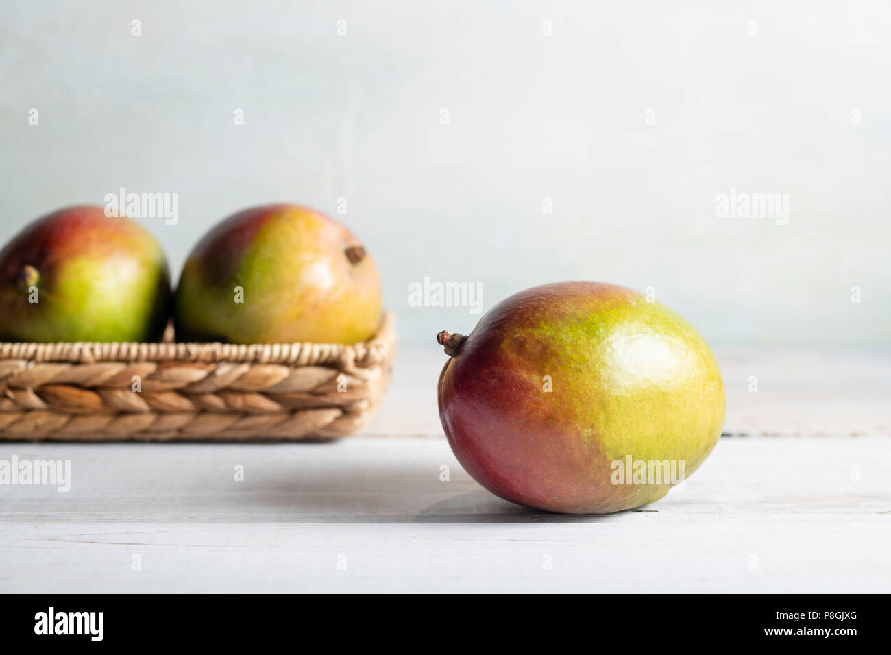 Drei reife Mangos in einem Weidenkorb auf ein whitewash Holz Tisch mit hellen, Tageslicht. Stockfoto