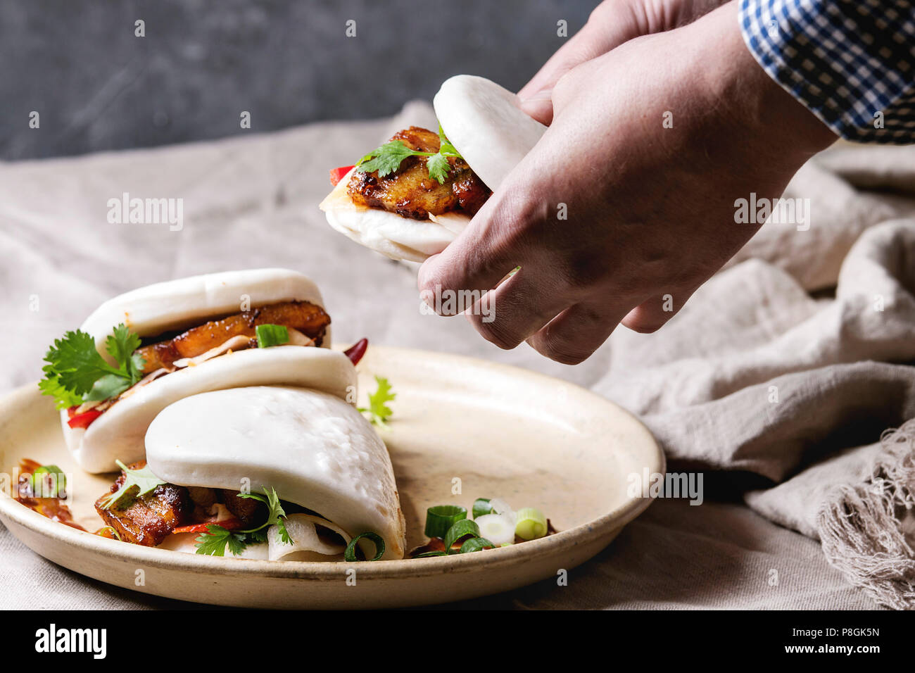 Des Menschen Hände halten asiatische Sandwich gedämpft Gua bao Brötchen mit Schweinebauch, grünen und Gemüse in Keramik Teller am Tisch serviert mit Bettwäsche Tischdecke. Wie Stockfoto