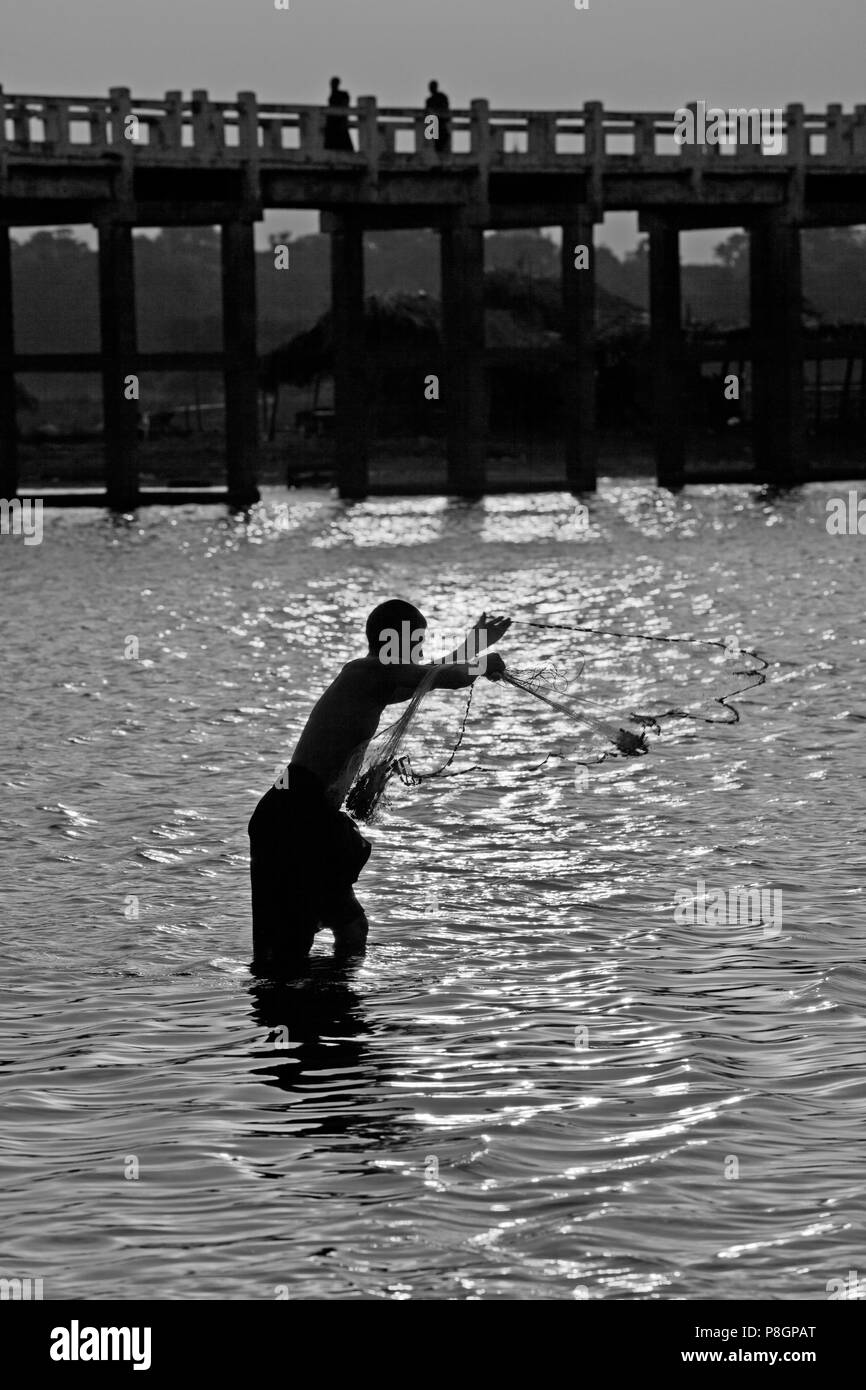 Ein Fischer wirft sein Netz unter U KLANGKUNST Brücke auf Taungthaman See bei Sonnenaufgang - AMARAPURA, MYANMAR Stockfoto