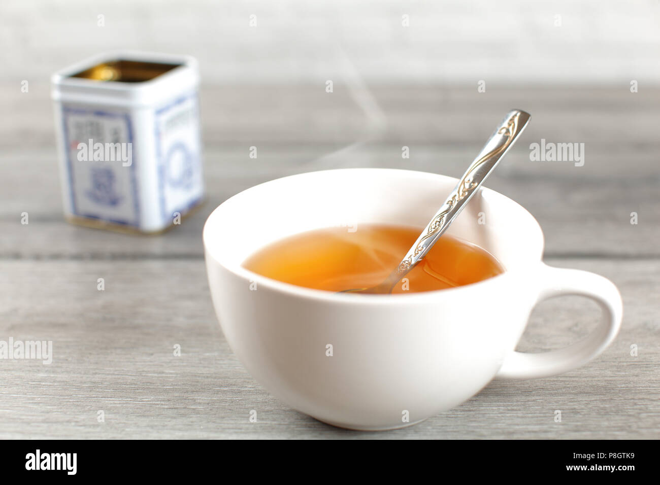 Heiße Tasse gelb Tee mit Dampf, mit Metall Zinn auf grau Holz Schreibtisch im Hintergrund platziert. Stockfoto