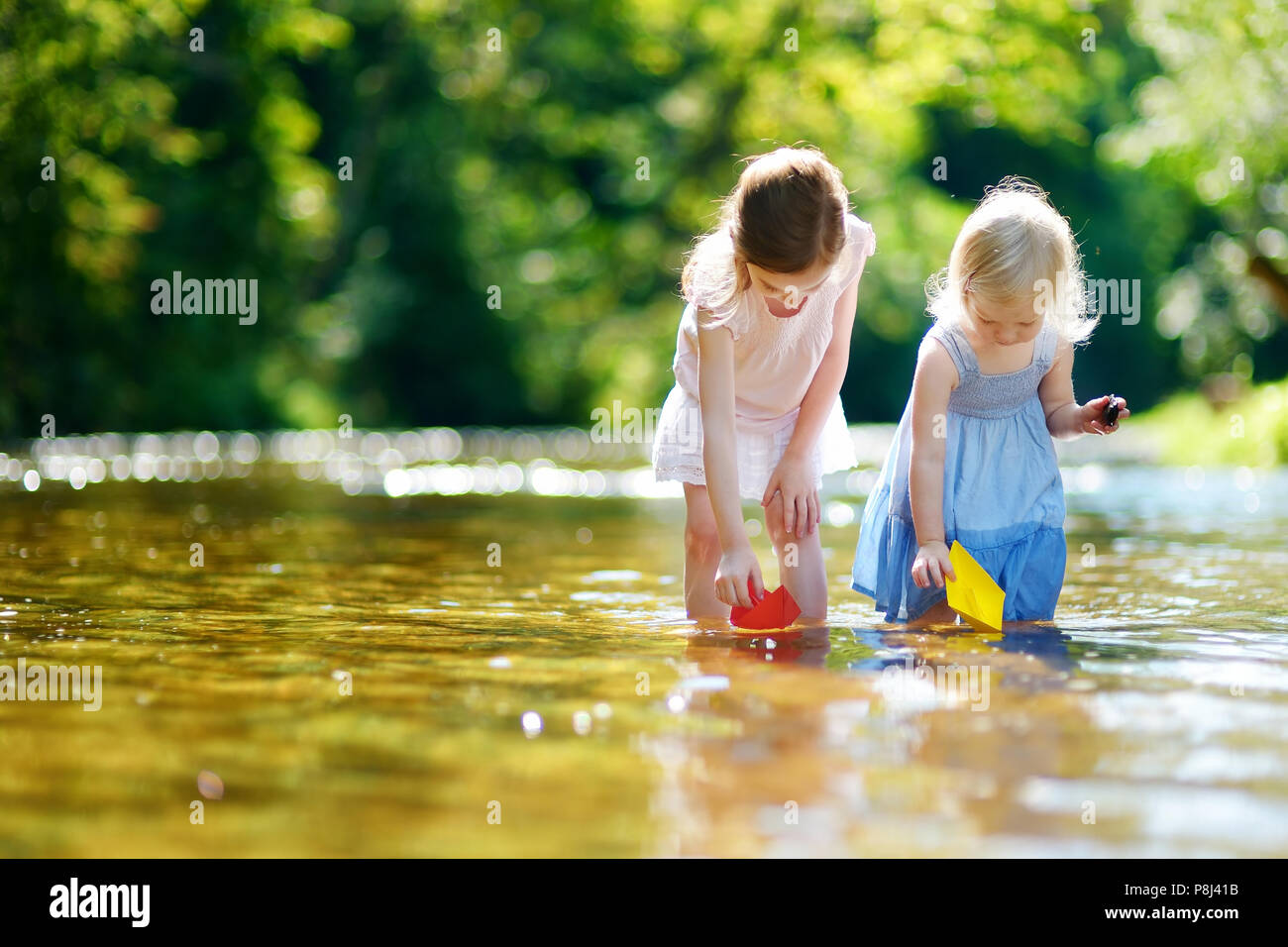 Zwei Entzückende kleine Schwestern spielen mit Papier Boote durch einen Fluss an warmen und sonnigen Sommer Tag Stockfoto