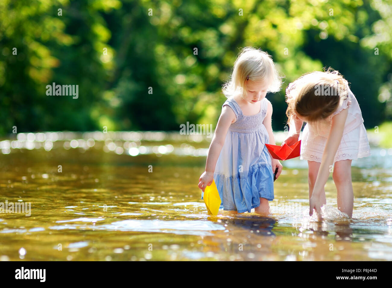 Zwei Entzückende kleine Schwestern spielen mit Papier Boote durch einen Fluss an warmen und sonnigen Sommer Tag Stockfoto