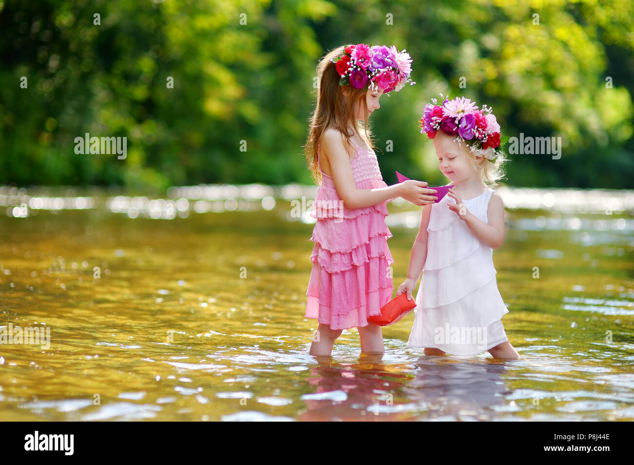 Zwei Entzückende kleine Schwestern tragen Blume Kronen spielen mit Papier Boote durch einen Fluss an warmen und sonnigen Sommer Tag Stockfoto
