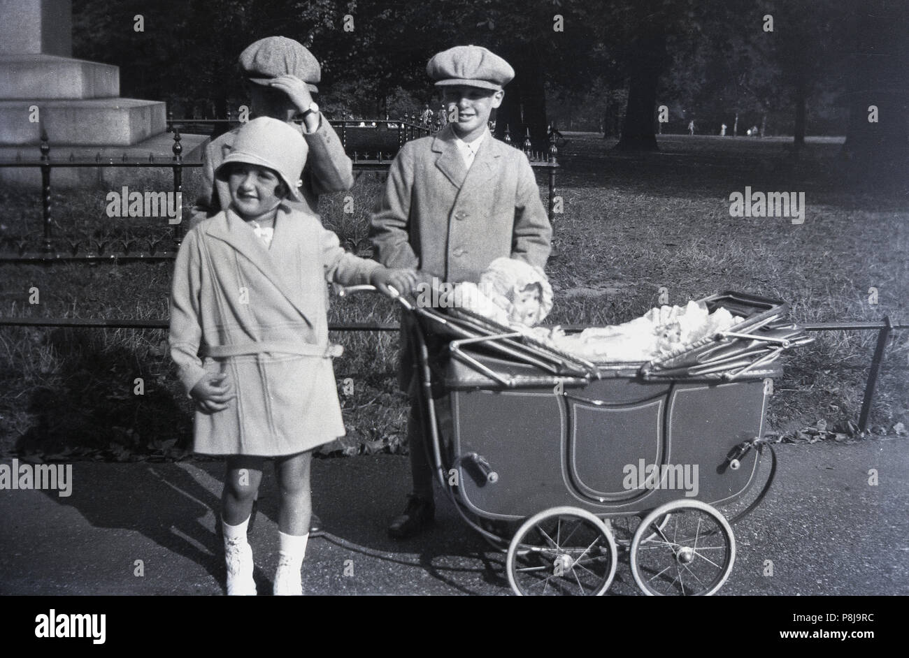 1920s, historische, drei elegant gekleidete Kinder, die von einem Park das Tragen der Kleidung des Tages - die beiden Jungs haben beide flacher Deckel Hüte auf", newsboy' mit Stil - mit den jungen Mädchen Stolz von pram ihre hübschen Puppen, England, UK. Stockfoto