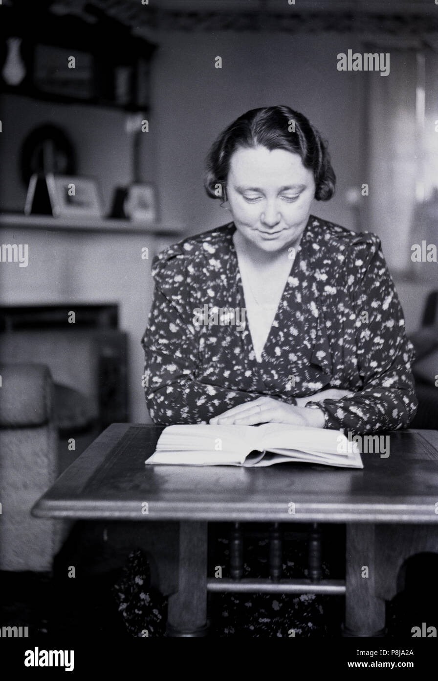 C, 1940er Jahre, historische, eine Dame trägt ein gemustertes Kleid an einem kleinen Tisch sitzen in einem Zimmer vorne ein Buch lesen, England, UK. Stockfoto
