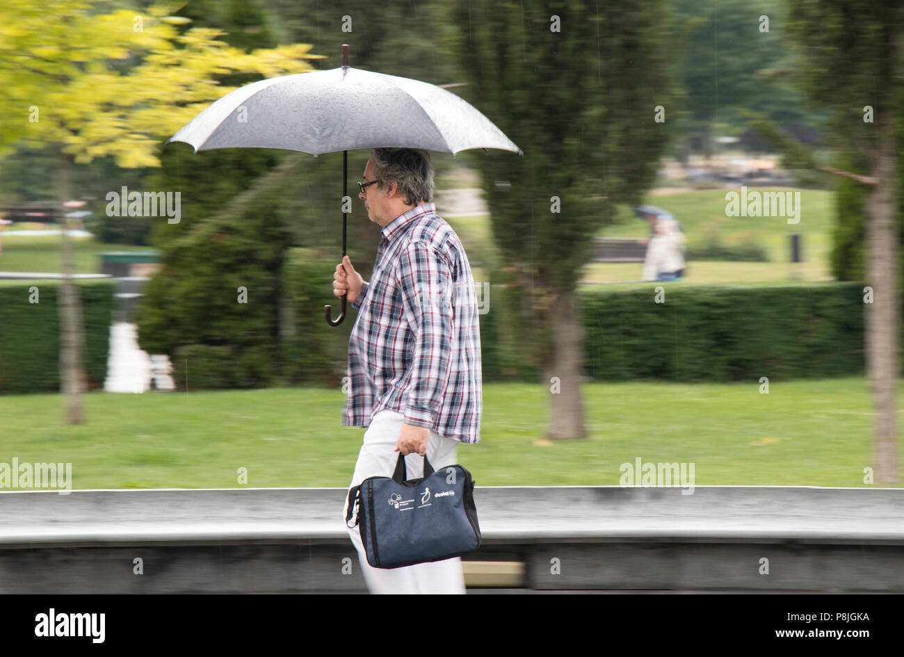 Belgrad, Serbien - Juni 14, 2018: Ein mittleres Alter Mann unter dem Dach im Frühjahr Regen im Stadtpark, im Besitz einer Tasche Stockfoto