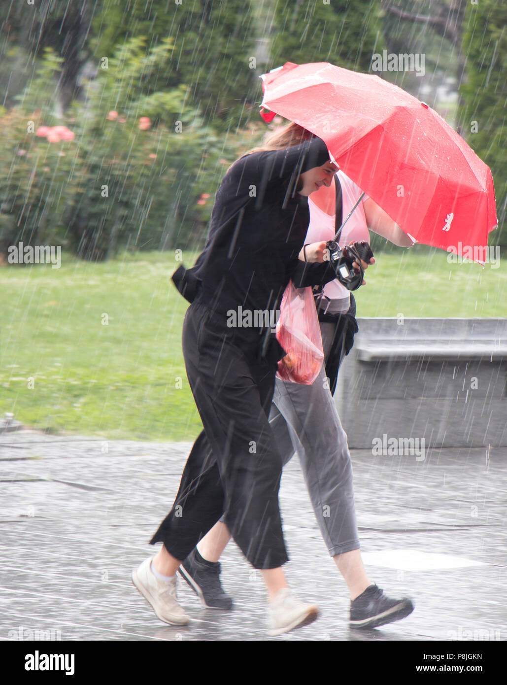 Belgrad, Serbien - Juni 14, 2018: Zwei junge Frauen unter roten Regenschirm in die plötzliche schwere Feder Regen in der Stadt Park läuft, Halten einer Kamera und Stockfoto