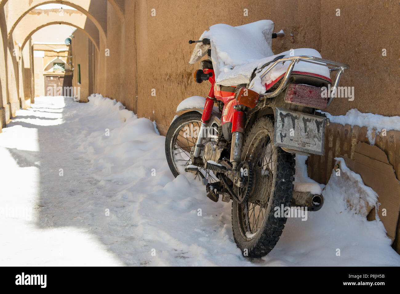 Eine verschneite Motorrad ruht gegen eine Lehm und Stroh Wand in einem Schnee erzeugt zurück Passage in der Innenstadt von Yazd, Iran gefüllt. Stockfoto