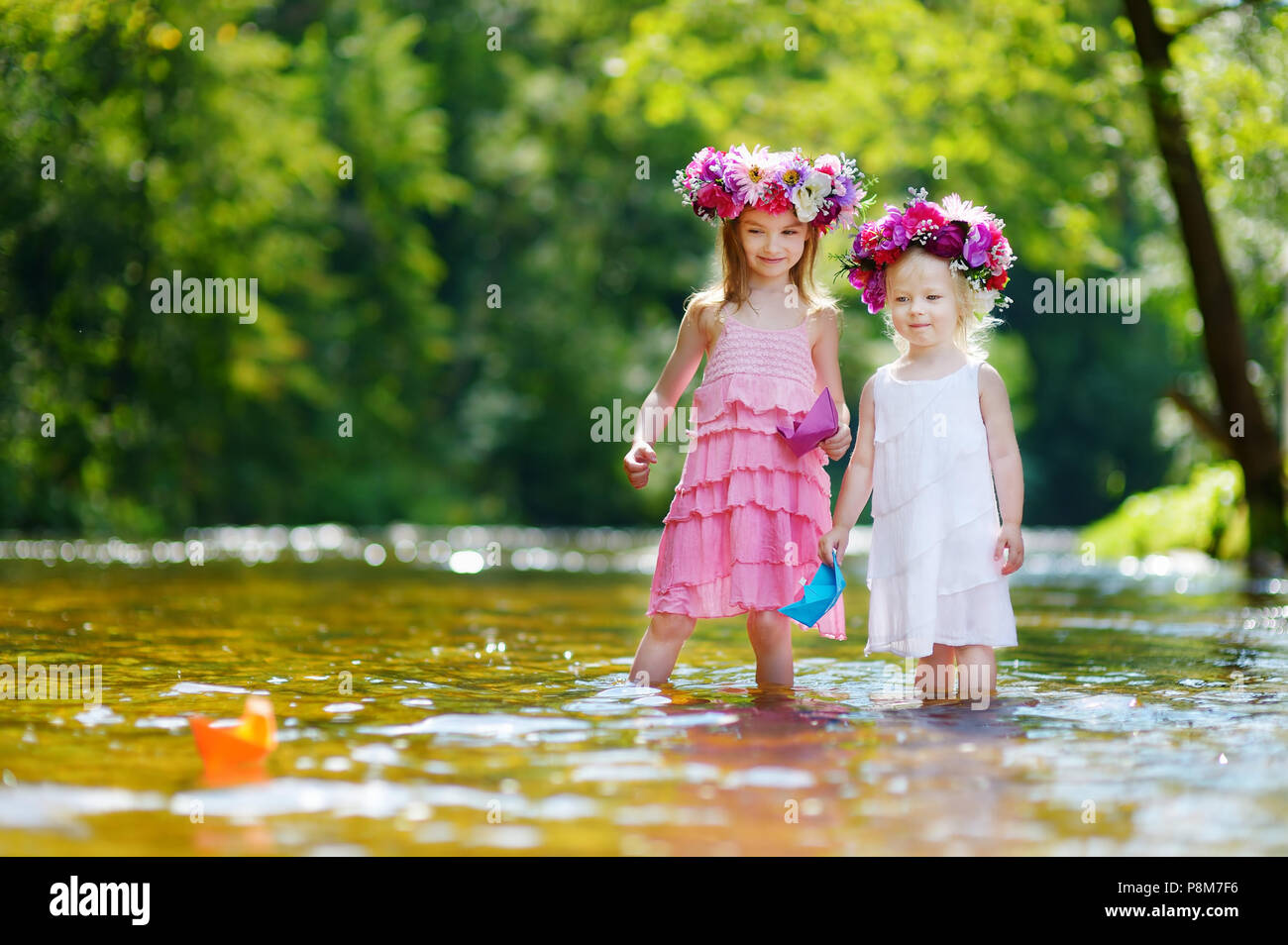 Zwei Entzückende kleine Schwestern tragen Blume Kronen spielen mit Papier Boote durch einen Fluss an warmen und sonnigen Sommer Tag Stockfoto