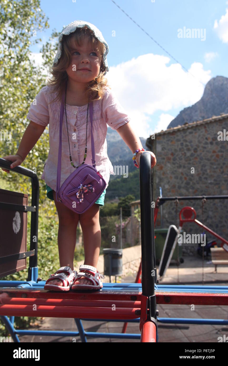 Schöne junge Kleinkind Mädchen in einem Play Park auf einem Klettergerüst mit einem lila Handtasche in einer Motorhaube lächelnd Stockfoto