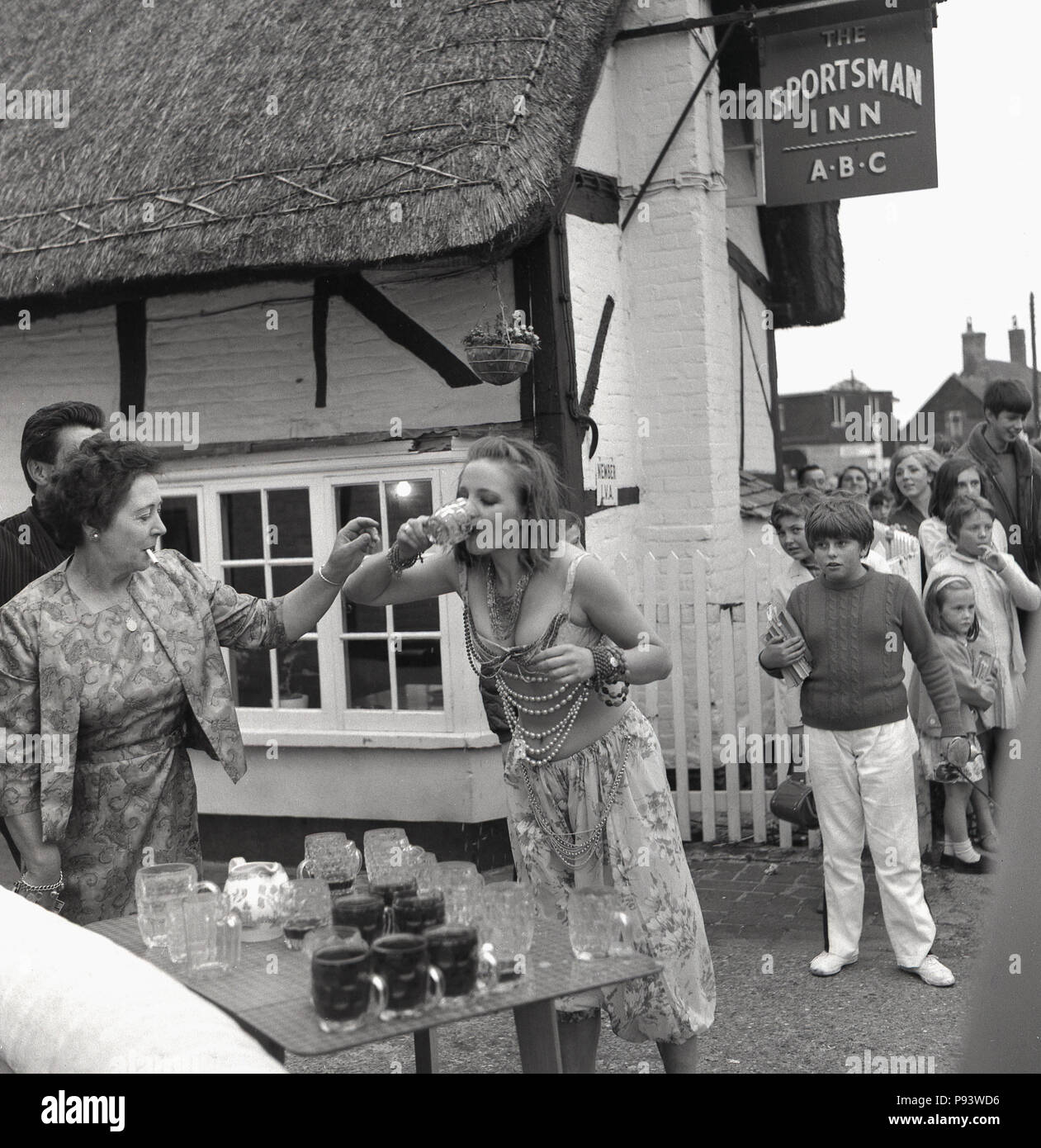 1967, eine junge Dame Fancy Dress schnell tragen Getränke ein Glas Bier aus einer Tabelle von leeren und vollen Bierkrügen außerhalb der Sportsman Inn in Quainton Dorf, Aylesbury, Bucks, England, UK. Mit fag im Mund, die elegant gekleideten Wirtin ist zur Hand, um das Glas zu erheben, während eine Gruppe von Jugendlichen auf Staunend betrachten. Die junge Dame ist konkurrieren in einem trinkendes Spiel als Teil der Dorffest feiern. Stockfoto