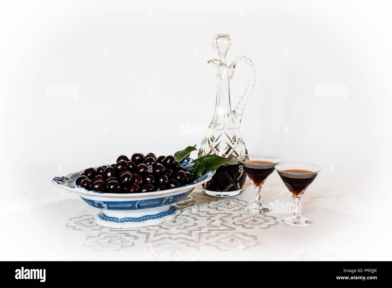 Kirschen auf einem alten China Teller und einen Kristall Karaffe und Gläser mit Cherry Brandy auf einem Tuch mit einem weißen Hintergrund, ein Stilleben. Stockfoto