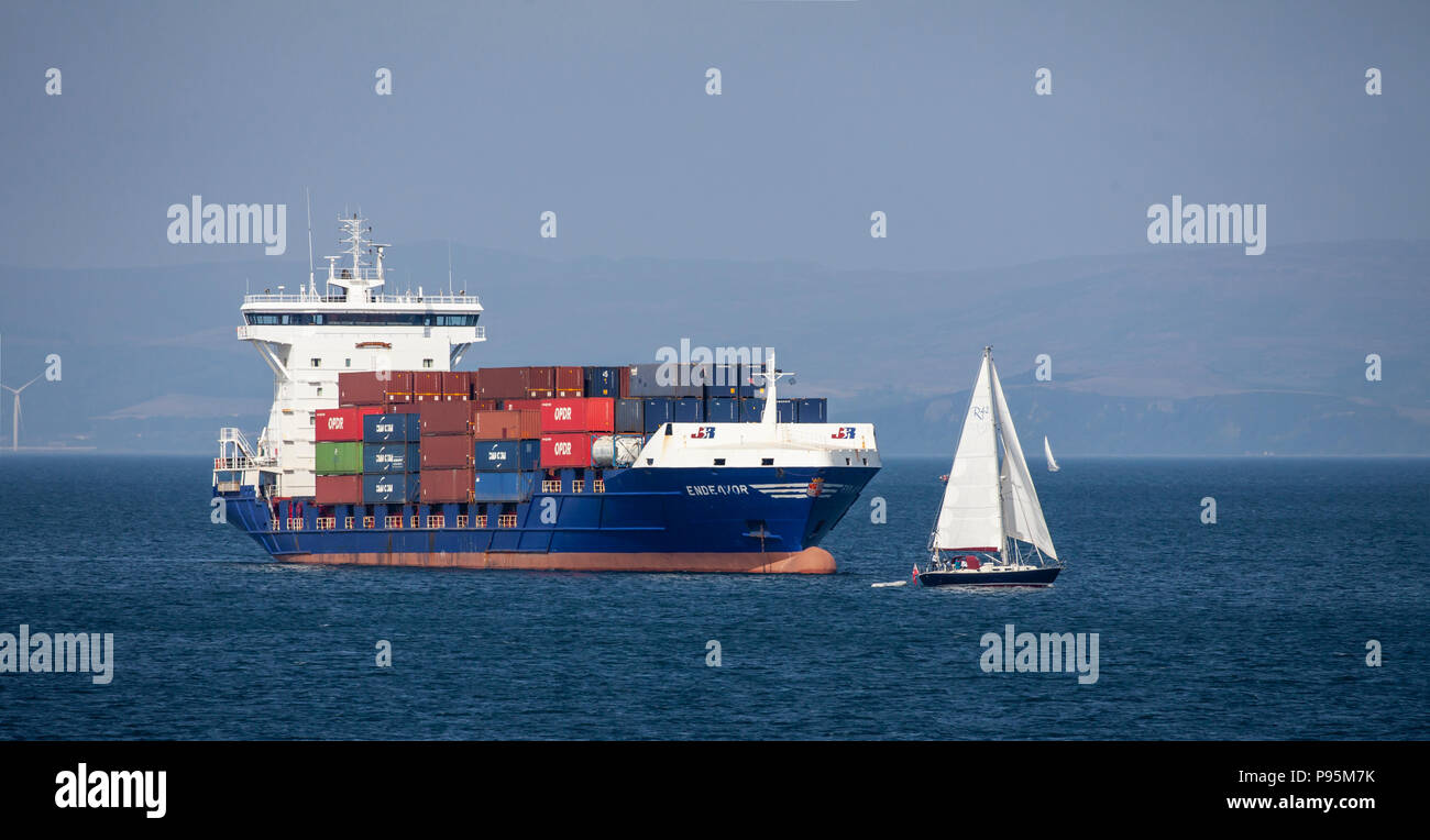 Das Container Feeder Ship MV Endeavour, im Besitz der niederländischen Firma JR Shipping, verpackt mit Containern, vor Anker in Brodick Bay, als eine Yacht vorbeifährt. Stockfoto