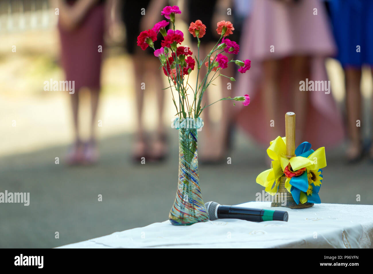 Blumenstrauß aus rosa und rote Blumen, Mikrofon und mit blauen und gelben Bändern Glocke auf unscharfen Personen silhouette sonnigen Hintergrund eingerichtet. Abschied Stockfoto