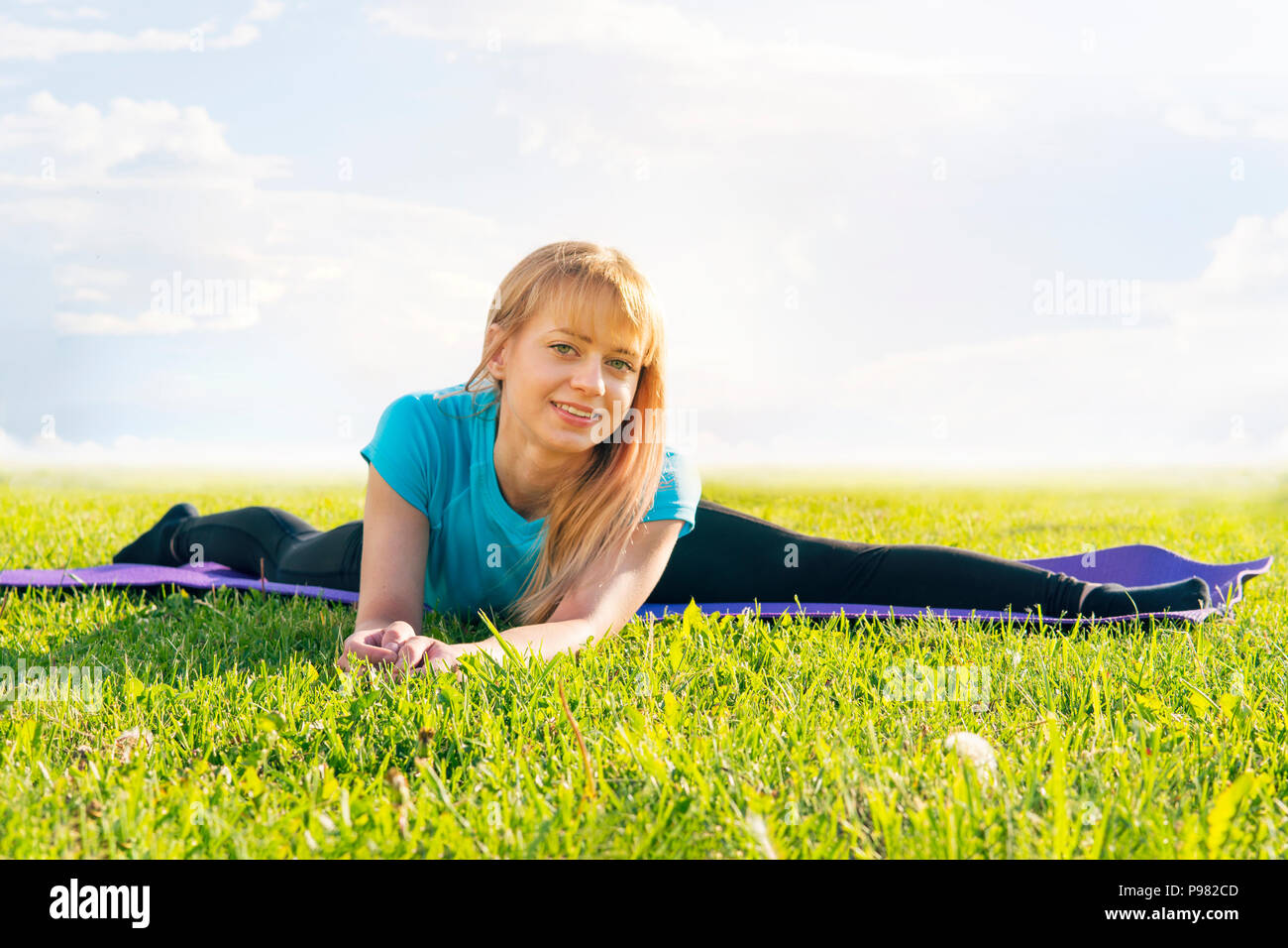 Eine blonde Frau Trainer in einem sportlichen kurzen Top und Fitnessraum leggings macht eine breite Garn auf der Wolldecke für Yoga Hände werden nach oben gerichtet auf die Yogamatte angehoben, auf einem Summ Stockfoto