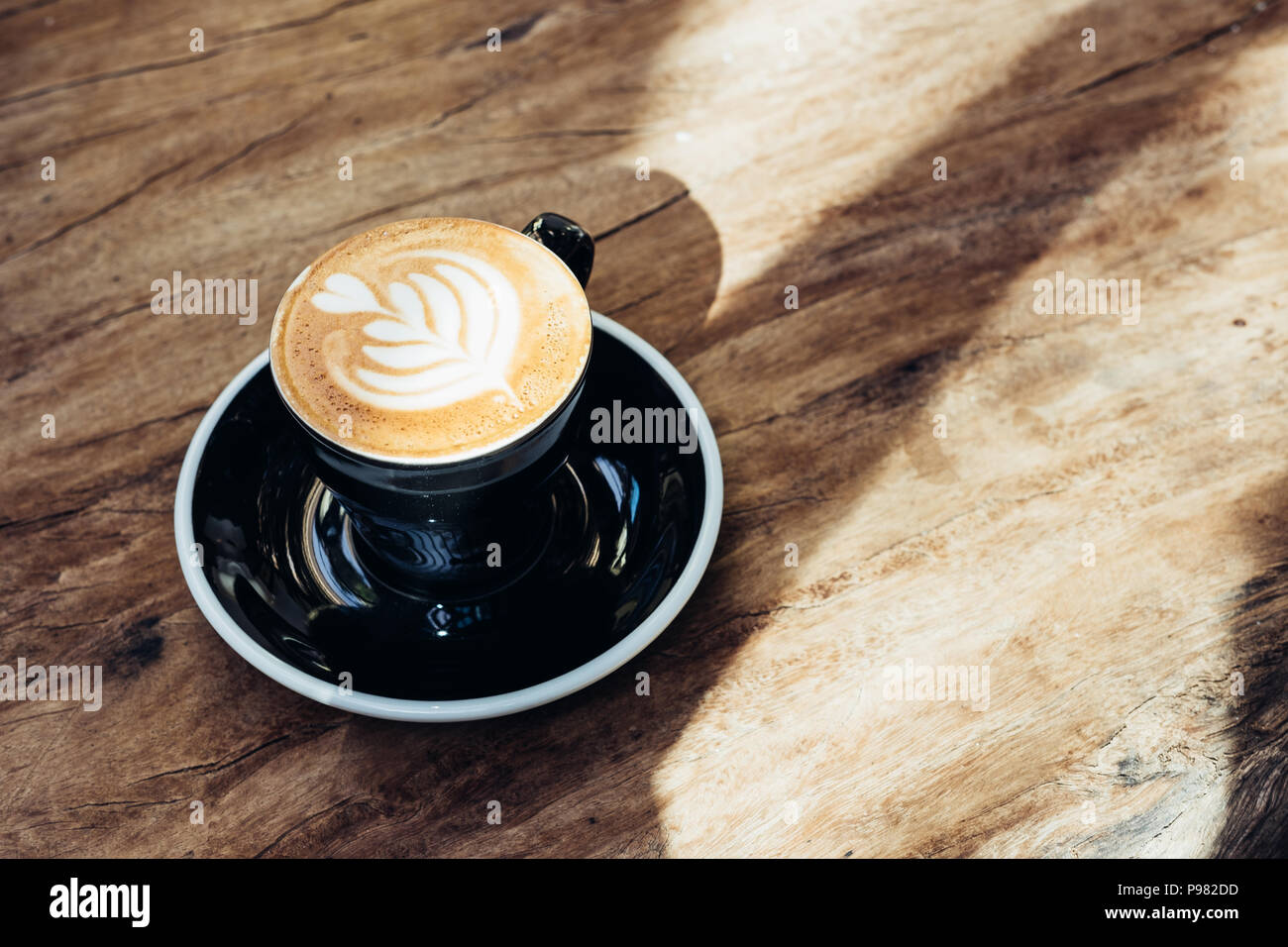 Close up Hot schwarz Kaffee Tasse mit Baum Herzform latte Art Milch Schaum auf Holz Tisch am Fenster mit harten Schatten Sonnenlicht im Cafe Restaurant. Stockfoto