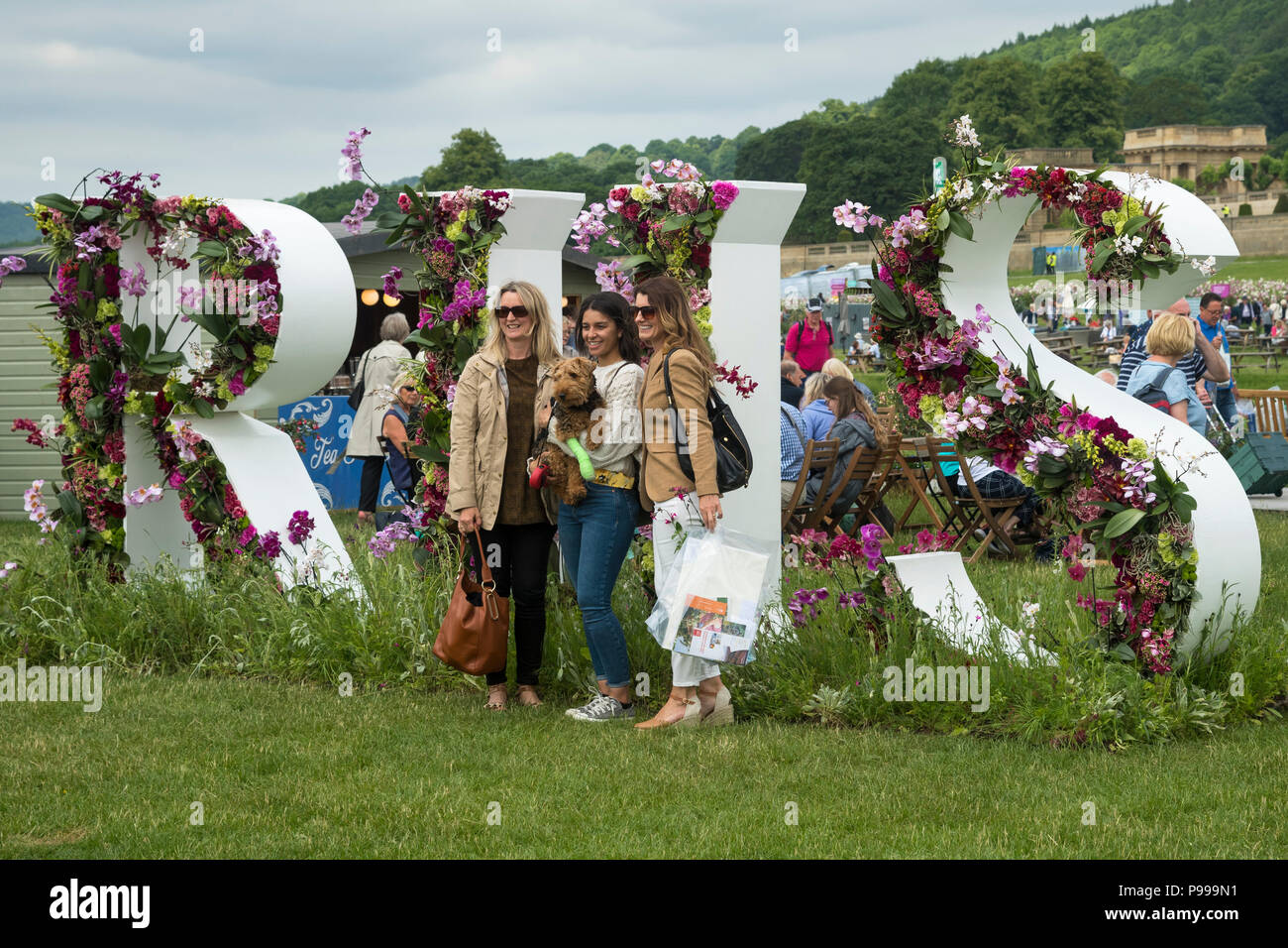 3 Frauen & Hund, lächelnd und für Fotos posiert, stehen vor großen Blumenarrangement (Buchstaben R H S) - Chatsworth Flower Show, Derbyshire, England, UK. Stockfoto