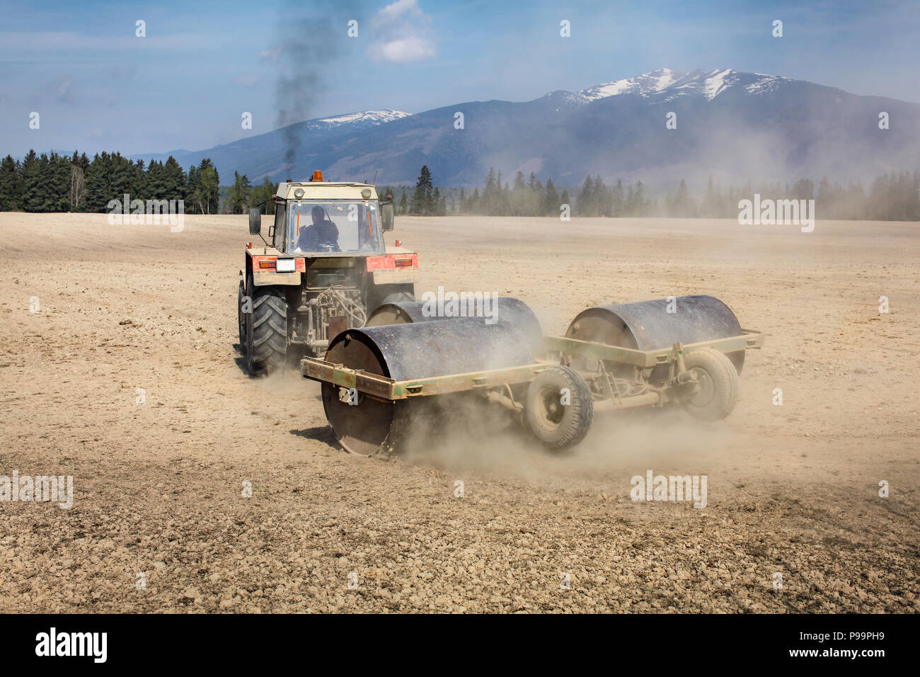 Tractor Pulling heavy metal Walze auf trockenen feld, Staub hinter und schwarzer Rauch oben, Berge im Rücken. Frühling Boden Vorbereitung. Stockfoto