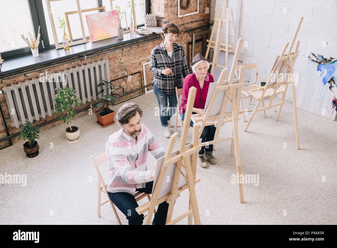 Hohen winkel Porträt von zwei erwachsenen Kursteilnehmer Malerei sitzt von staffeleien in art studio mit weiblichen kunst Lehrer beobachten Sie in geräumigen und lichtdurchfluteten Loft spac Stockfoto