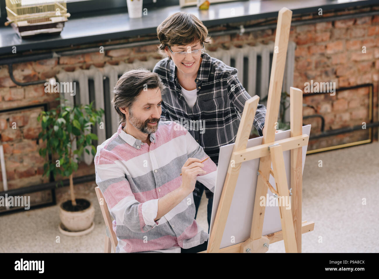 Hohen winkel Portrait von reifer Mann Gemälde von staffelei sitzen in art studio mit weiblichen kunst Lehrer ihn in der Kunst der Klasse, kopieren Raum Stockfoto