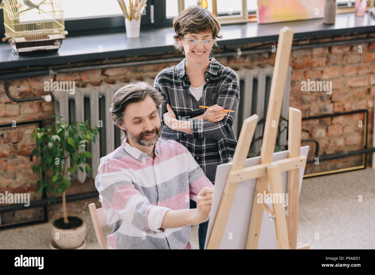 Portrait von lächelnden reifer Mann Malerei sitzend durch Staffelei im Atelier mit weiblichen kunst Lehrer ihn in der Kunst der Klasse, kopieren Raum Stockfoto