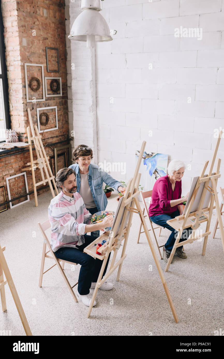 Hohen winkel Portrait von erwachsenen Kursteilnehmer Malerei sitzt von staffeleien in art studio mit weiblichen kunst Lehrer, kopieren Raum Stockfoto
