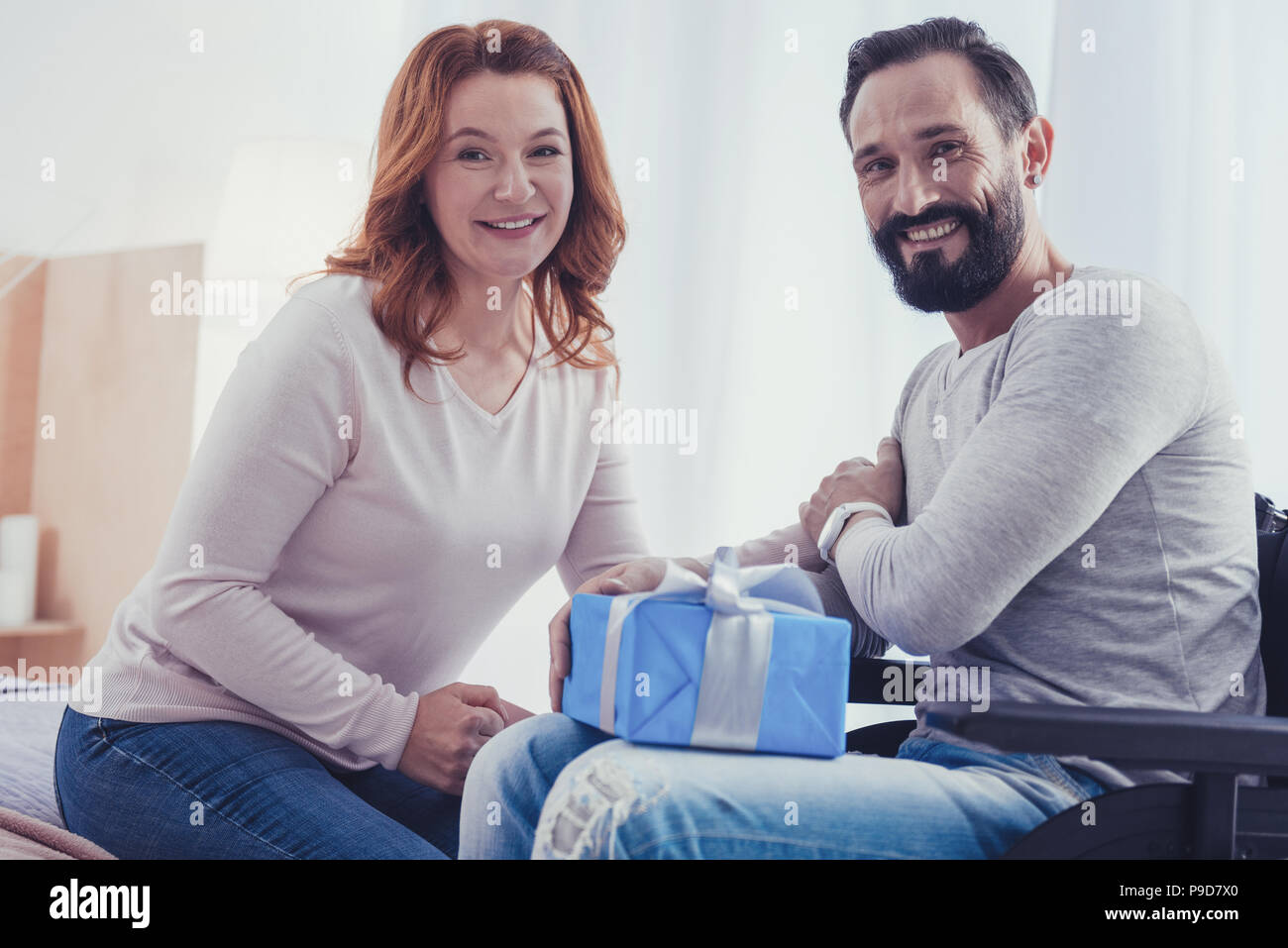 Behinderte Mann sitzt mit einem hübschen präsent, während seine Frau in der Nähe, und lächelnd Stockfoto