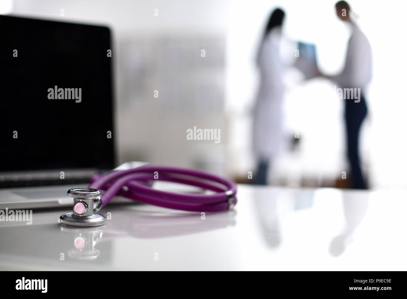Laptop und Stethoskop auf dem Schreibtisch, Ärzte im Hintergrund stehend Stockfoto